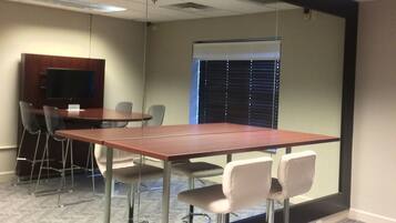 Een vergaderruimte met een lange tafel, vier stoelen, een projectiescherm en een raam met zonwering.