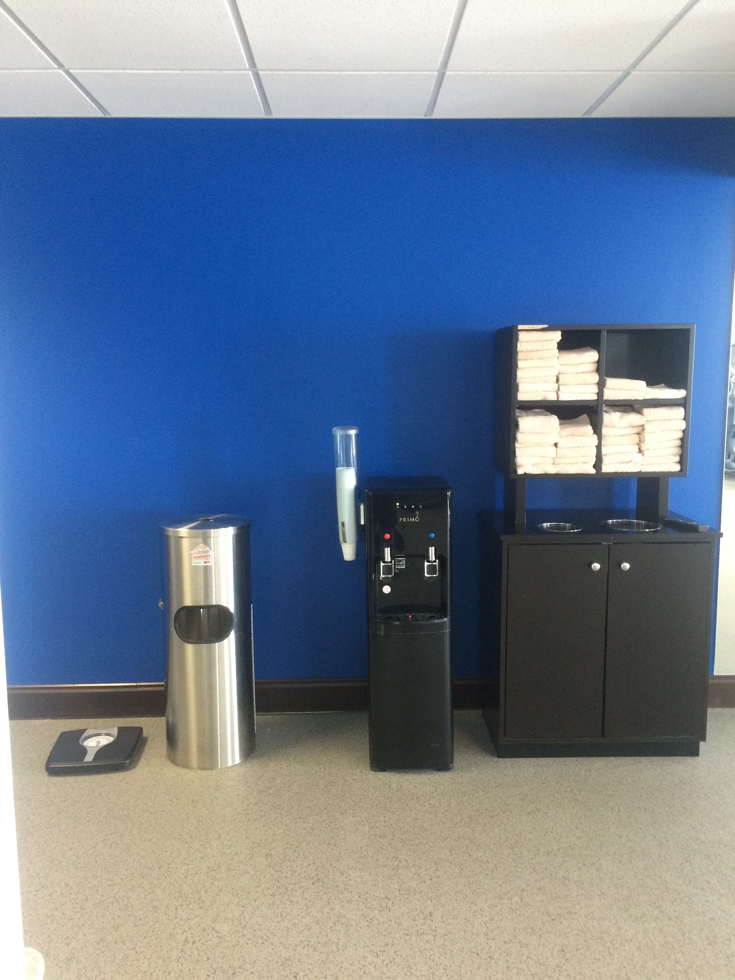 A blue wall, a trash can, a water dispenser, and a cabinet with folded towels.