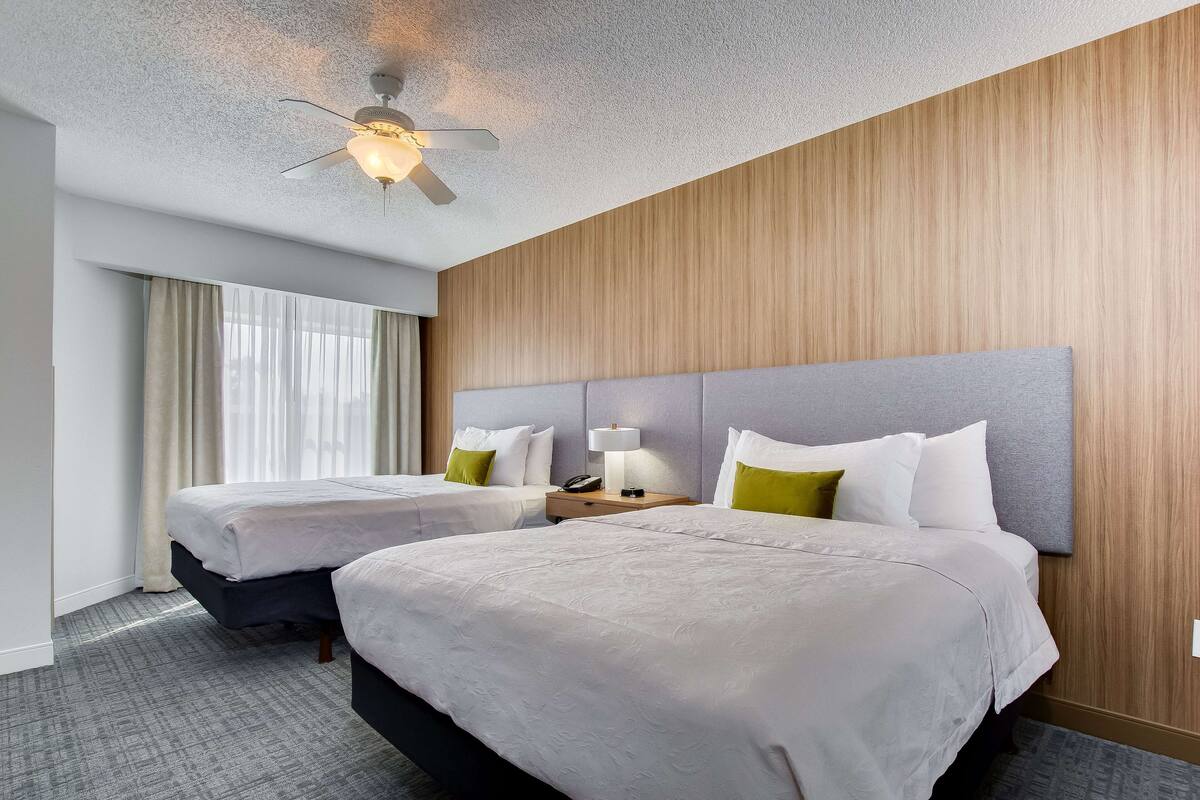 Camera d'albergo con due letti, una ventola a soffitto, un comodino con lampada e una finestra con tende.