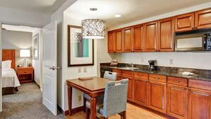 A hotel room with a wooden dining table, a chair, a bed, a nightstand, a lamp, and a kitchen area with wooden cabinets and a microwave. - Homewood Suites by Hilton Bentonville-Rogers (Rogers)