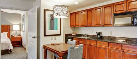 A hotel room with a wooden dining table, a chair, a bed, a nightstand, a lamp, and a kitchen area with wooden cabinets and a microwave.