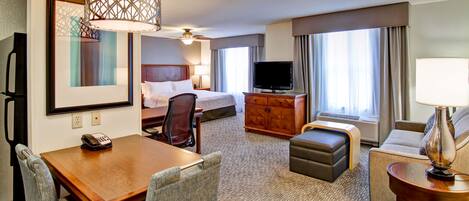 A hotel room with a bed, a TV, a desk with a telephone, a chair, a sofa, a lamp, and a large window with curtains.