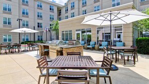 An outdoor patio area with multiple tables and chairs, a barbecue grill, and umbrellas providing shade. - Homewood Suites by Hilton Bentonville-Rogers (Rogers)