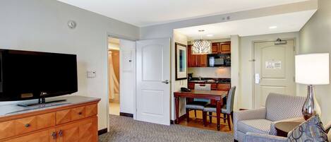 A hotel room with a flat-screen TV, wooden furniture, a dining area, and a kitchenette.