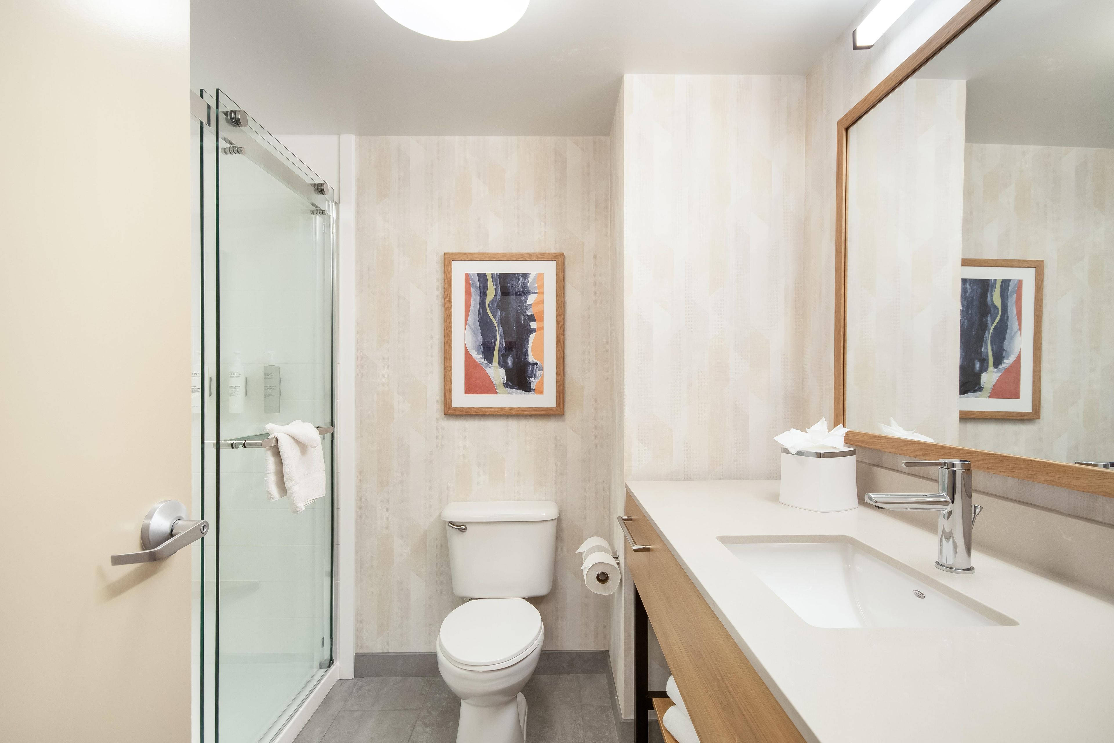 Un bagno moderno con box doccia in vetro, un water, un lavandino con specchio e un quadro astratto appeso alla parete.
