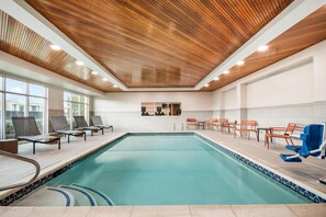 A swimming pool with lounge chairs and tables, wooden ceiling, and large windows.