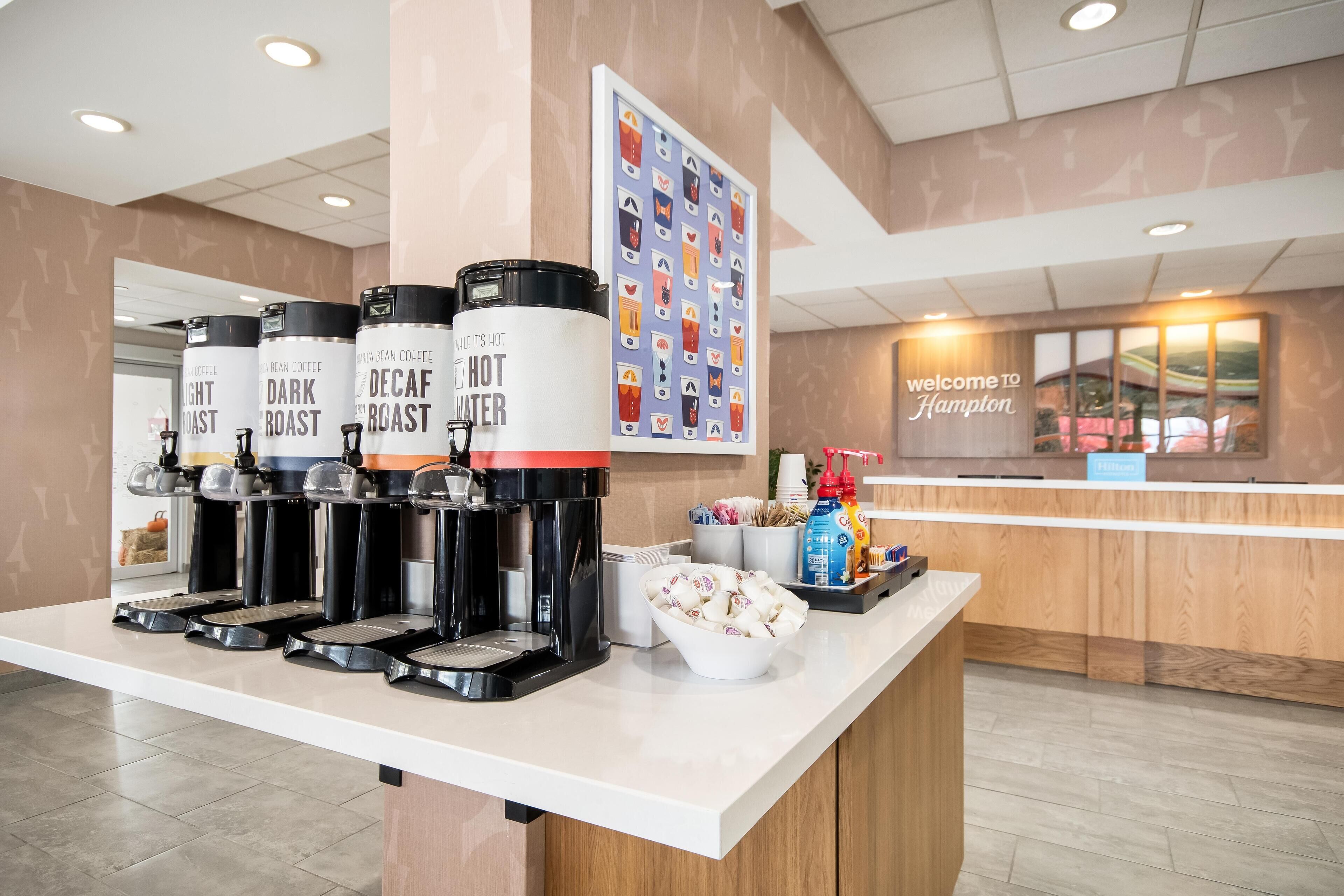 A coffee station with four dispensers labeled 'Dark Roast,' 'Light Roast,' 'Decaf,' and 'Hot Water.' There is a bowl of coffee pods and a 'Welcome to Hampton' sign in the background.
