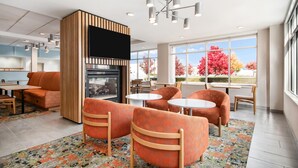 A modern lounge area with orange upholstered chairs, a fireplace, and large windows offering a view of autumn trees.