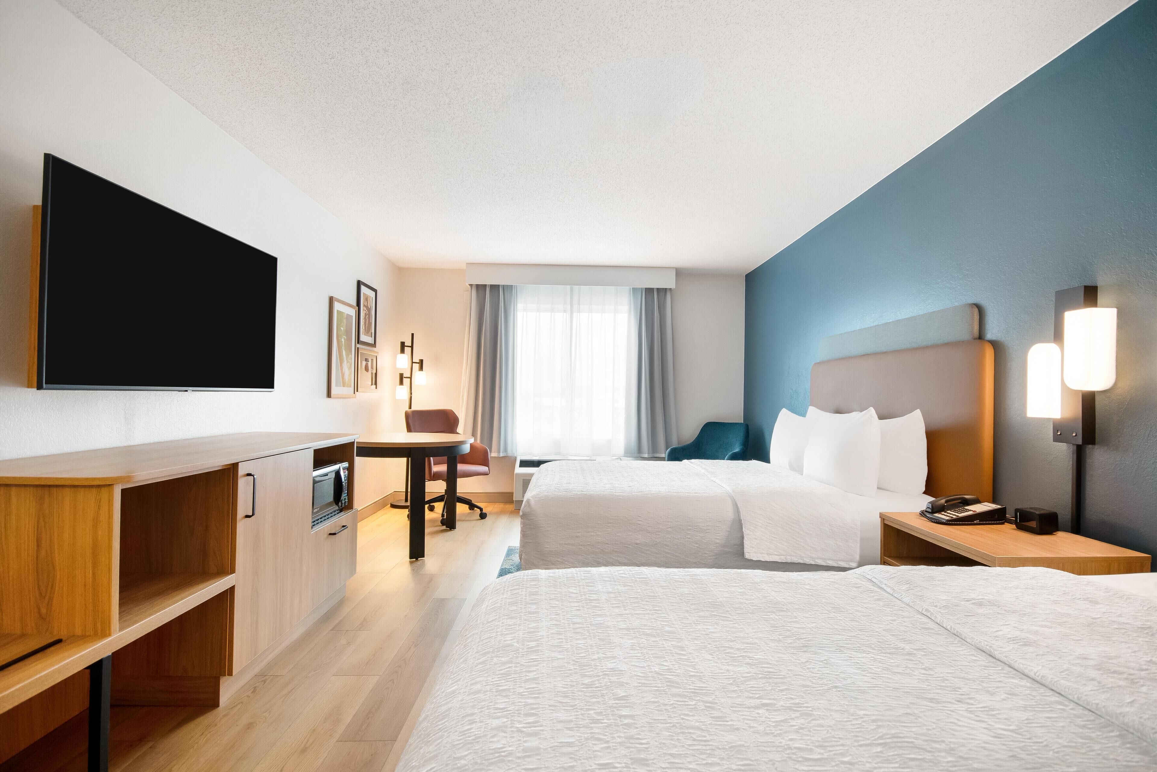 A hotel room with two beds, a desk, a flat-screen TV, a microwave, and a small table with a lamp.