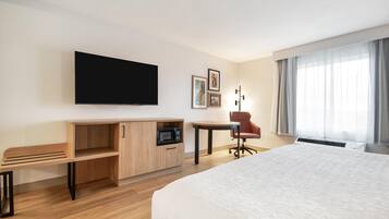 A hotel room with a bed, a desk with a microwave, a chair, a TV mounted on the wall, and a window with curtains.