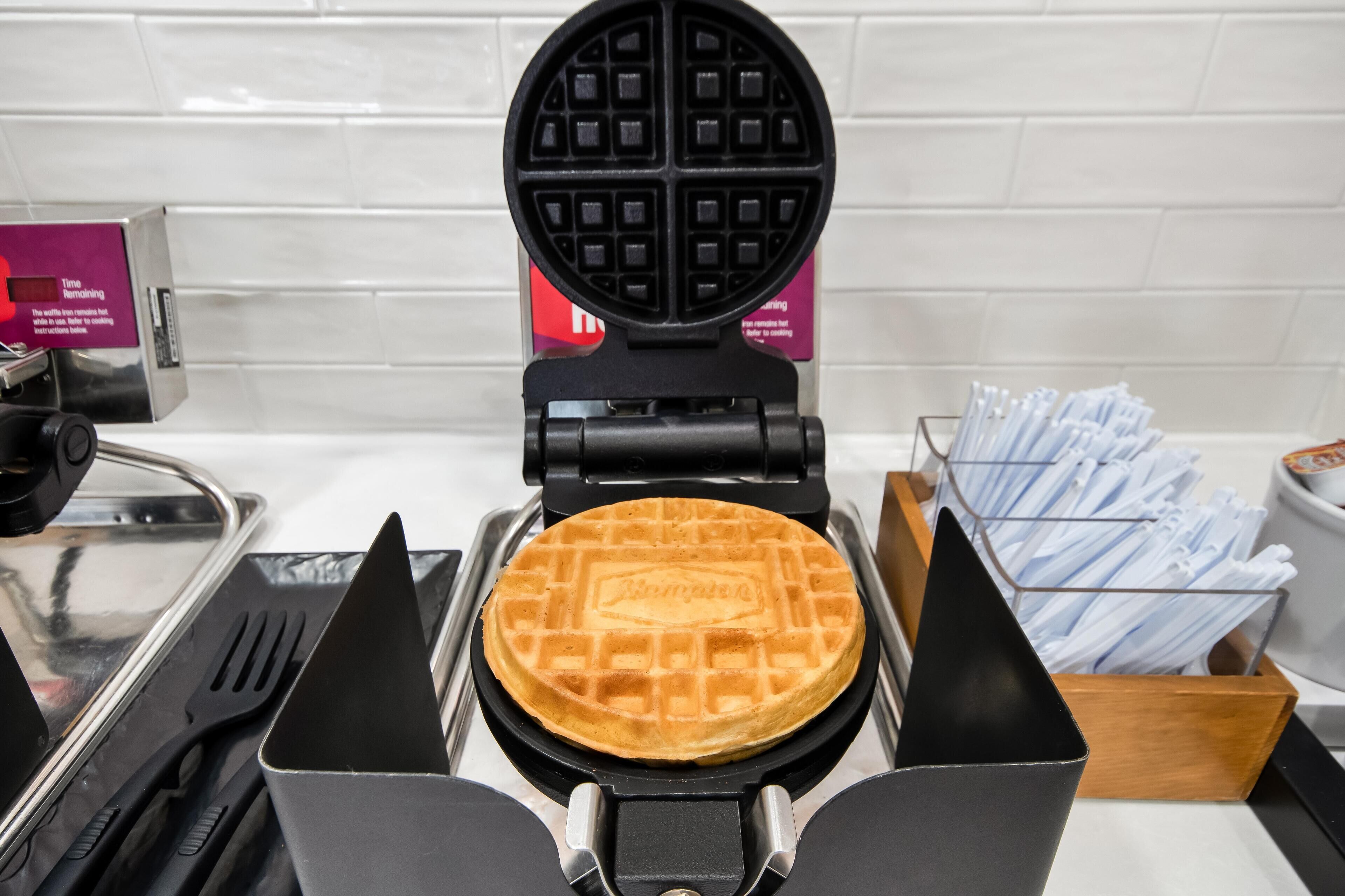 A waffle maker with a waffle inside, a spatula, a fork, and a box of napkins.