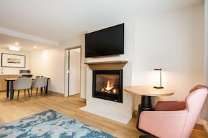 A modern living room with a fireplace, a flat-screen TV mounted above it, a dining area with chairs and a table, and a small round table with a lamp. - Hampton Inn Rutland (Rutland)