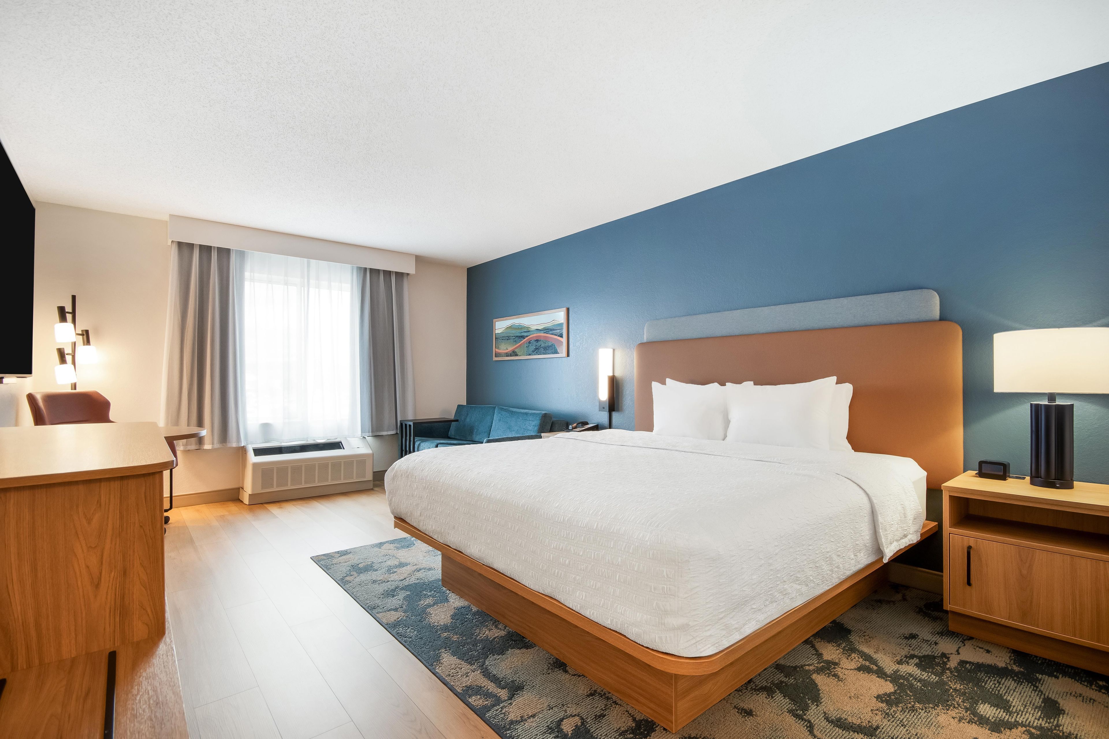 A hotel room with a large bed, a desk, a chair, a nightstand, a lamp, and a painting on the wall.