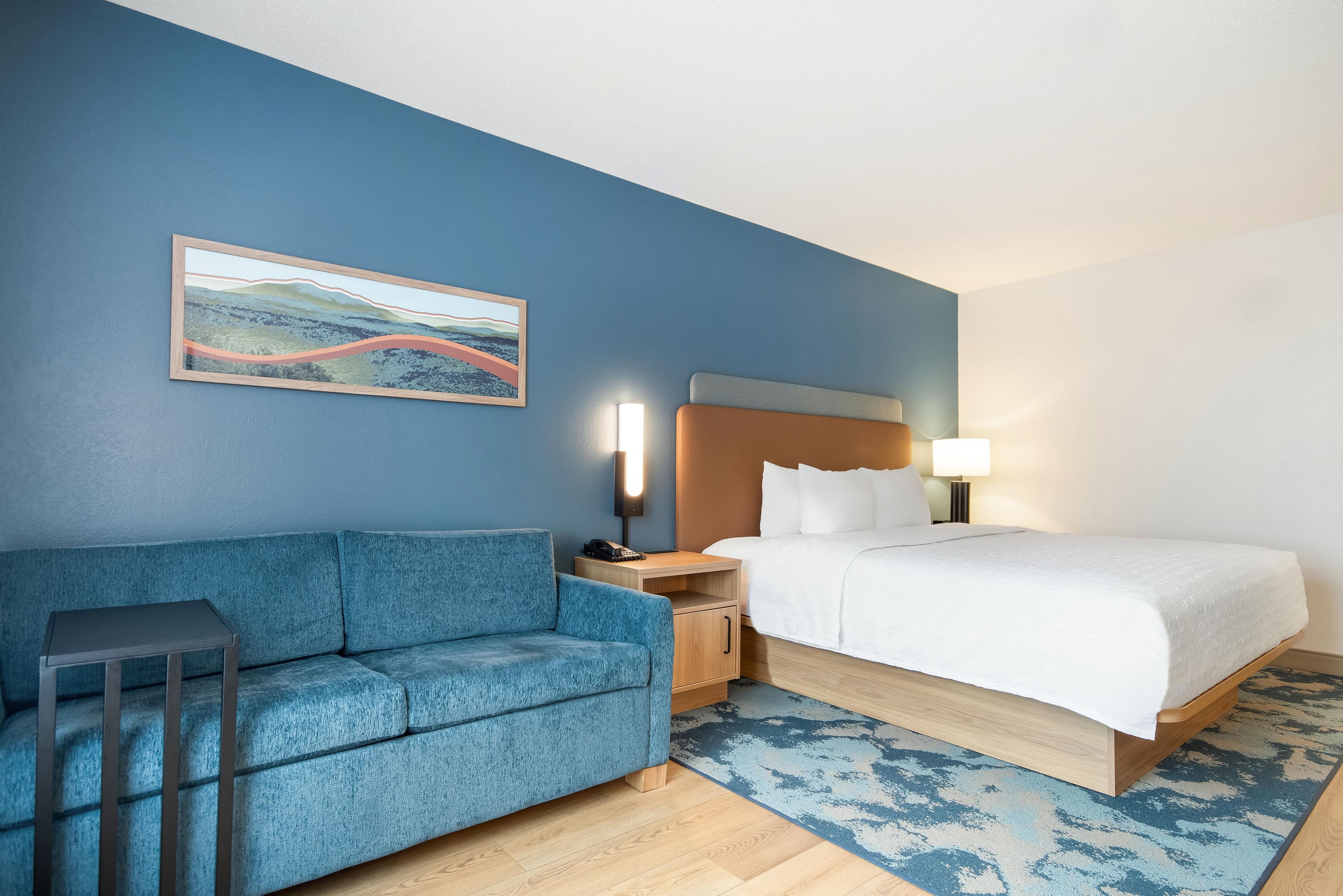 Una camera d'albergo con un divano blu, un letto con lenzuola bianche, un comodino con una lampada e un quadro appeso al muro.
