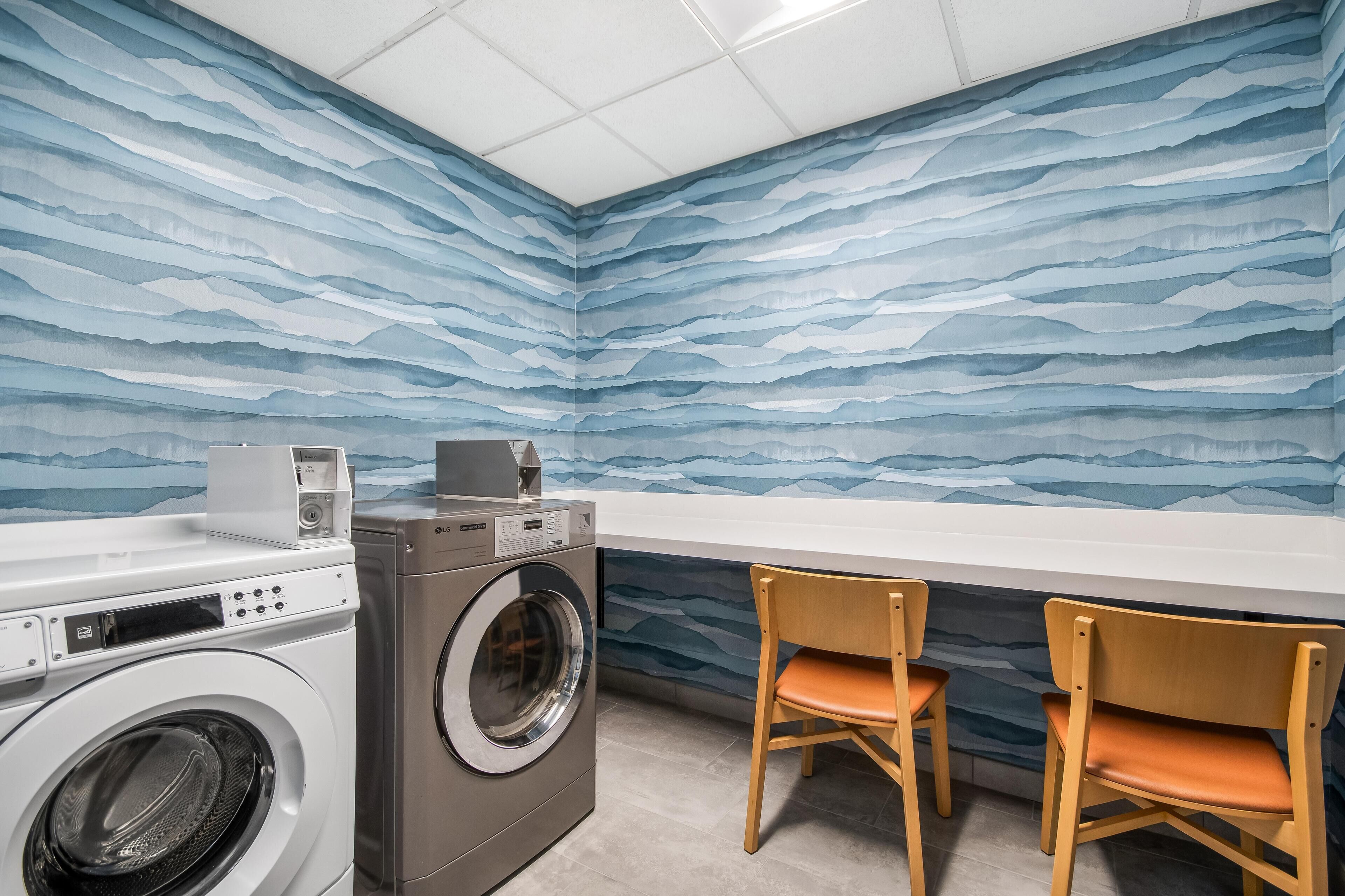 A laundry room with a washing machine, dryer, and two wooden chairs.