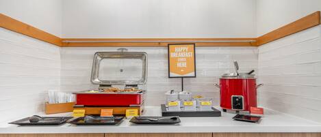 A breakfast station with a sign that reads "HAPPY BREAKFAST STARTS HERE," a hot food dispenser, a red slow cooker, and labeled containers for different food items.