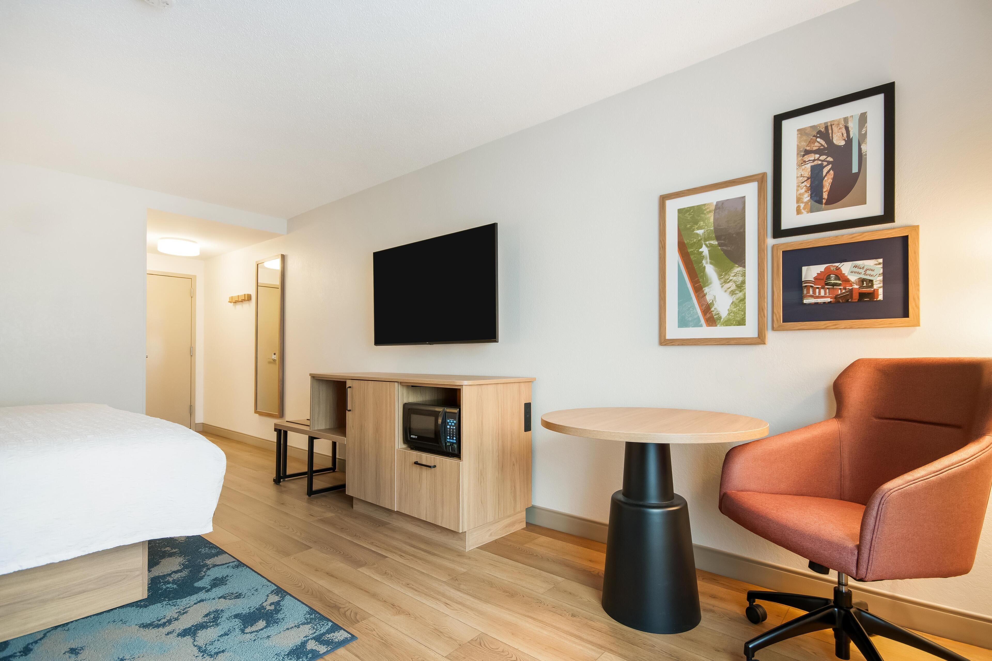 Una habitación de hotel con una cama, un escritorio con microondas, una silla, un televisor y cuadros en la pared.