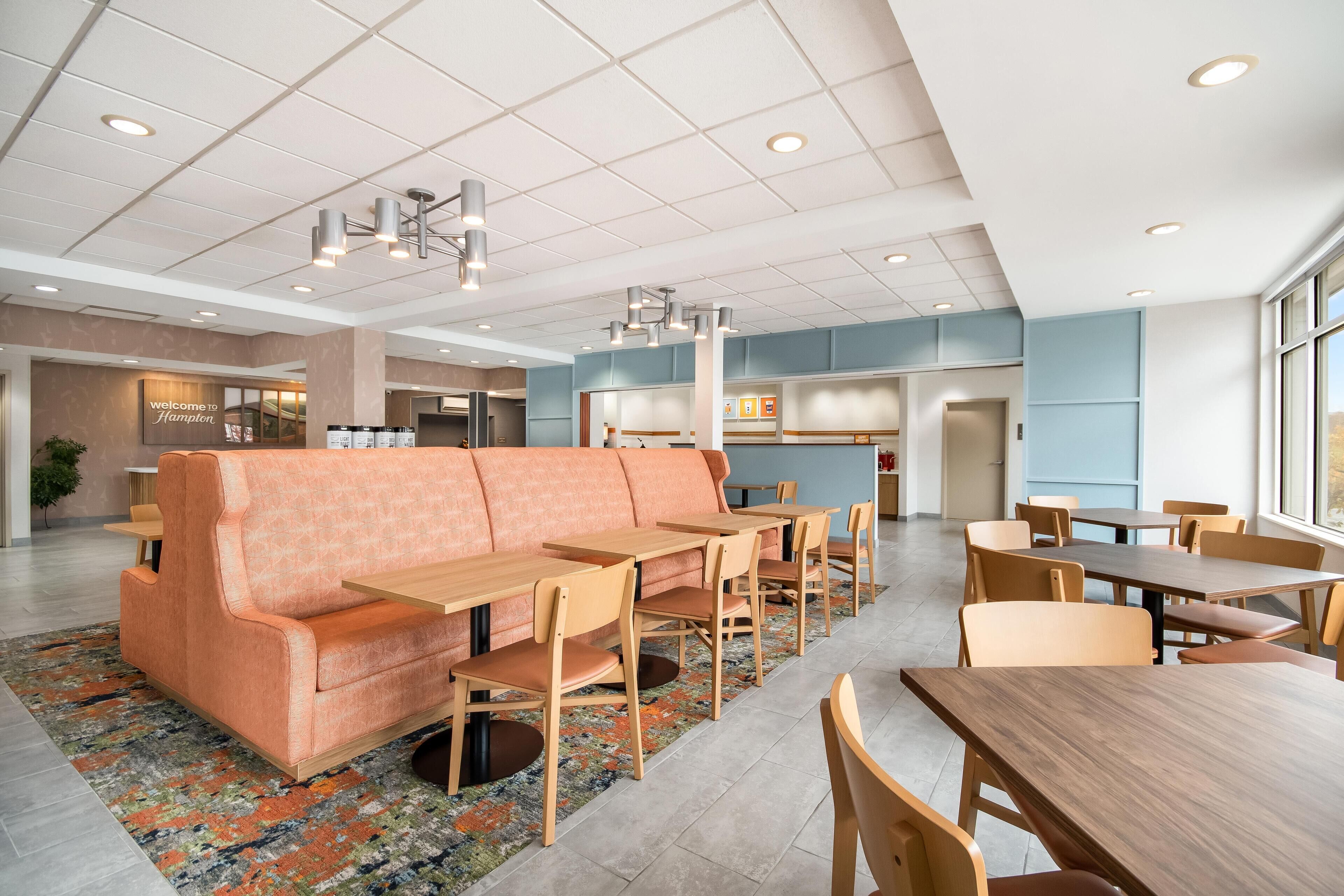 A modern hotel lobby with a seating area featuring a sofa and tables, a reception desk, and a large window providing natural light.