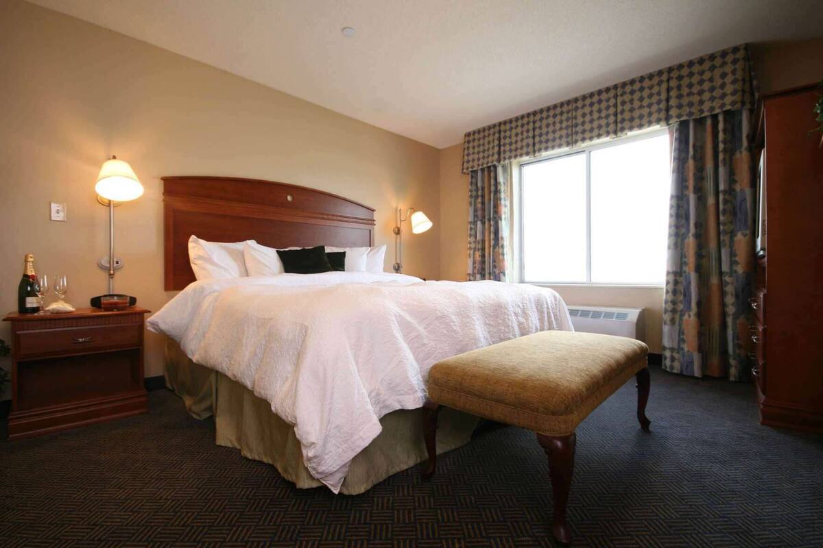 A hotel room with a large bed, bedside lamps, a nightstand, a window with curtains, and a carpeted floor.