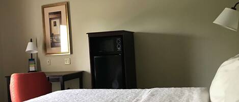 Una habitación de hotel con cama, escritorio con lámpara, refrigerador y un cuadro en la pared.
