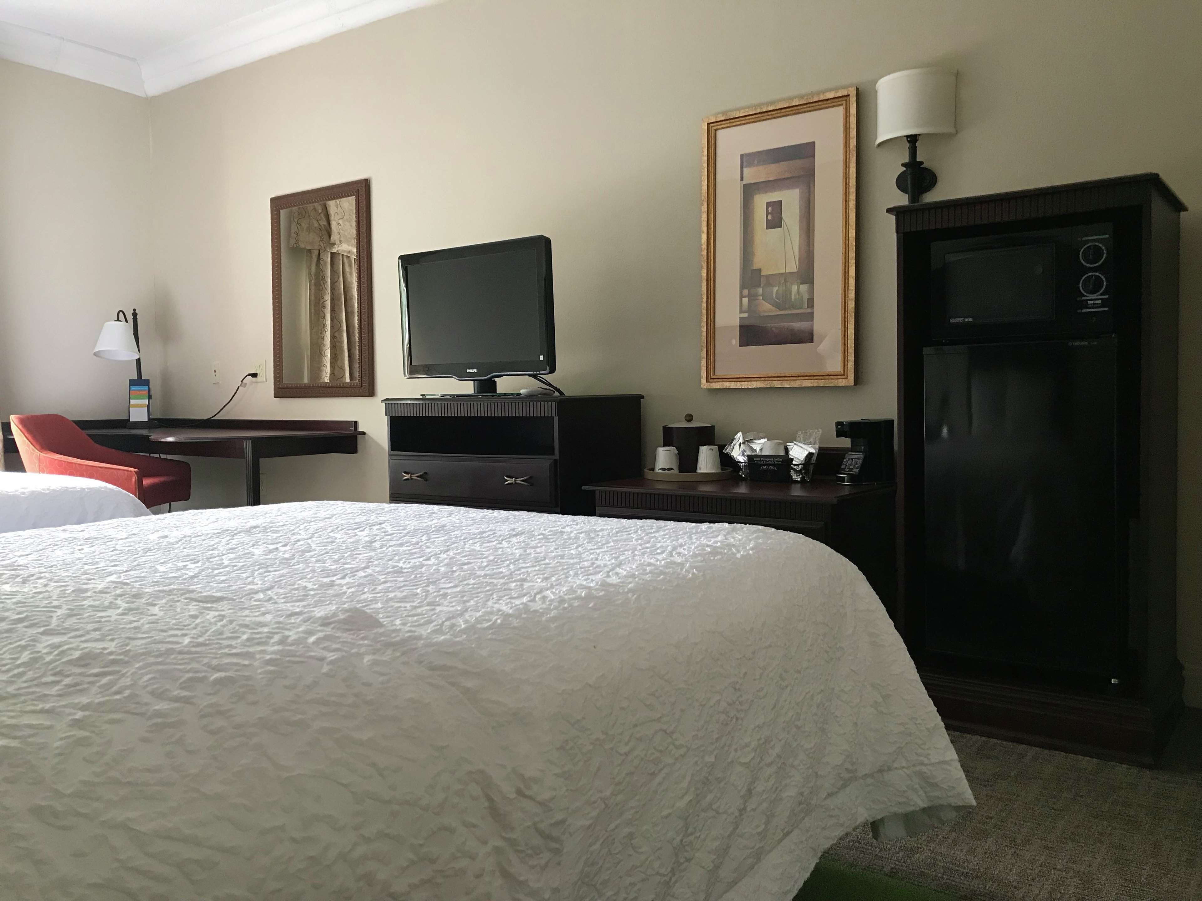 Habitación de hotel con una cama grande, un escritorio con computadora, televisor, refrigerador y cafetera.