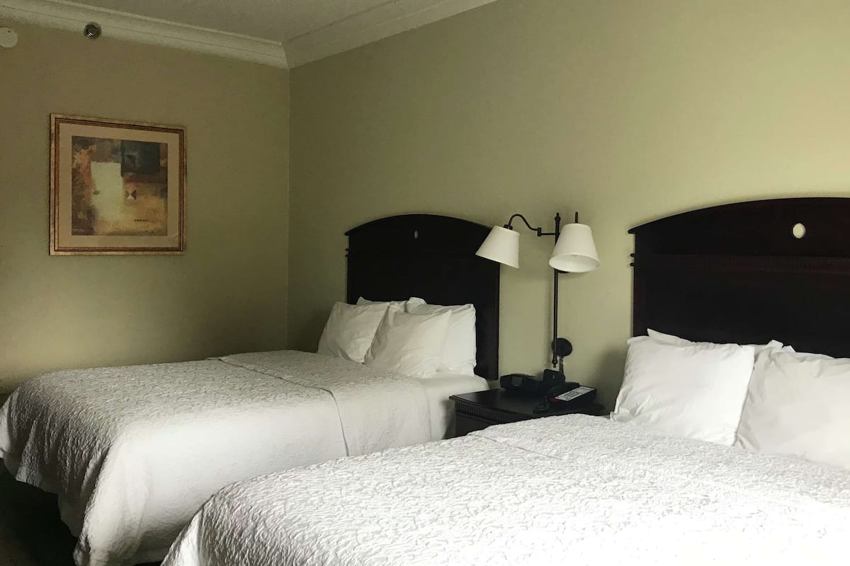 A hotel room with two single beds, a nightstand with a lamp, a painting on the wall, and a plain light-colored wall.