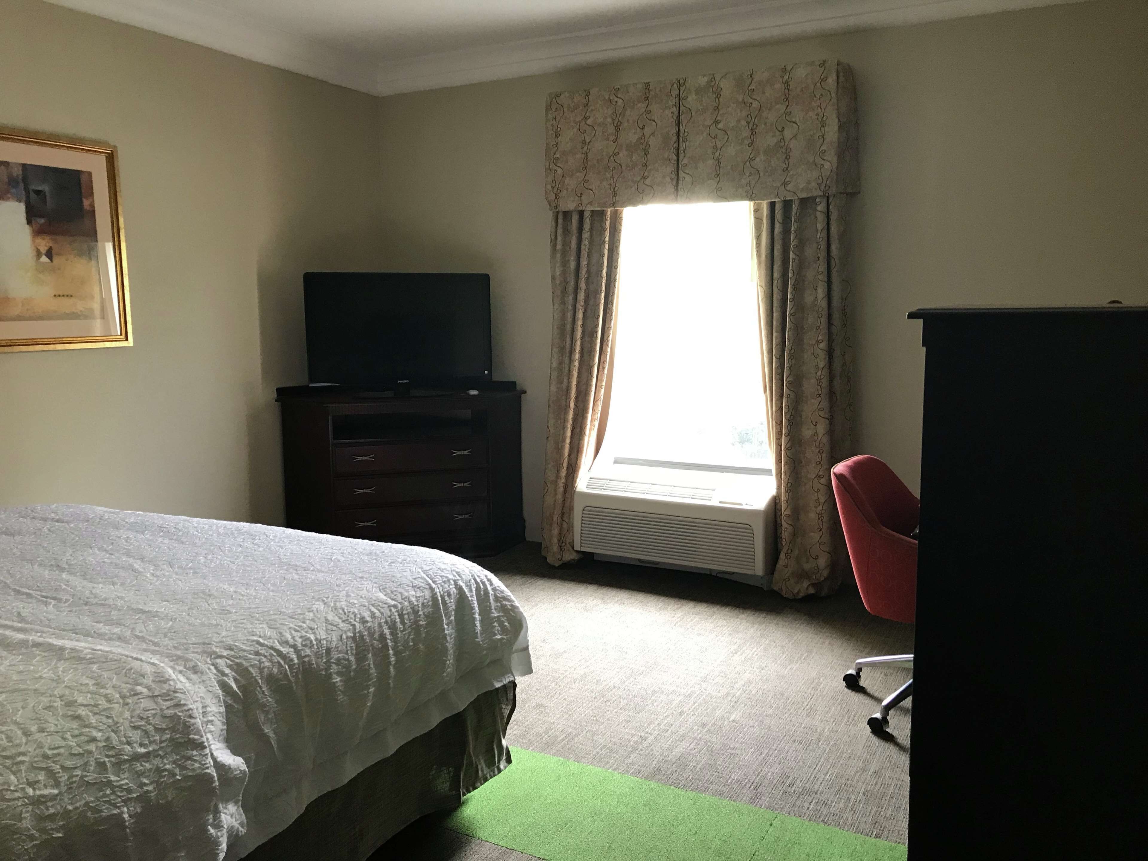 A hotel room with a bed, a television on a dresser, a desk with a chair, and a window with curtains.