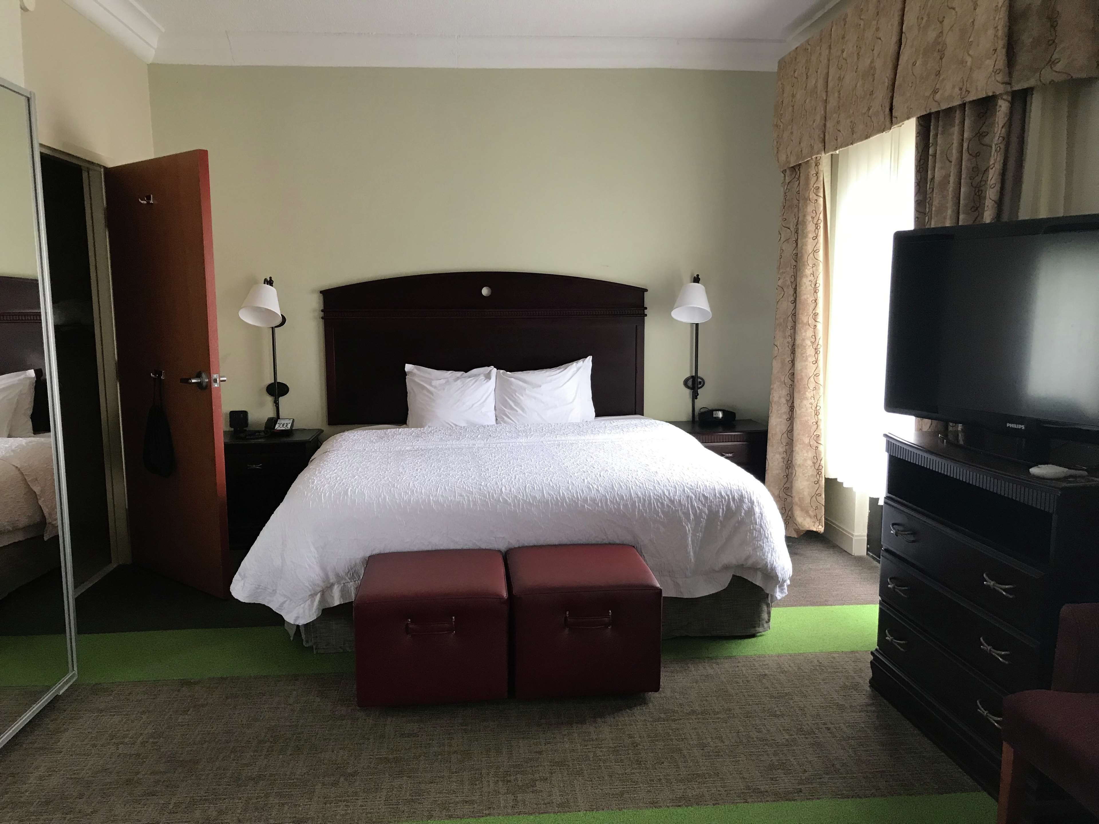 Quarto de hotel com uma cama grande, duas mesas de cabeceira com abajures, uma televisão, um guarda-roupa e um tapete verde.