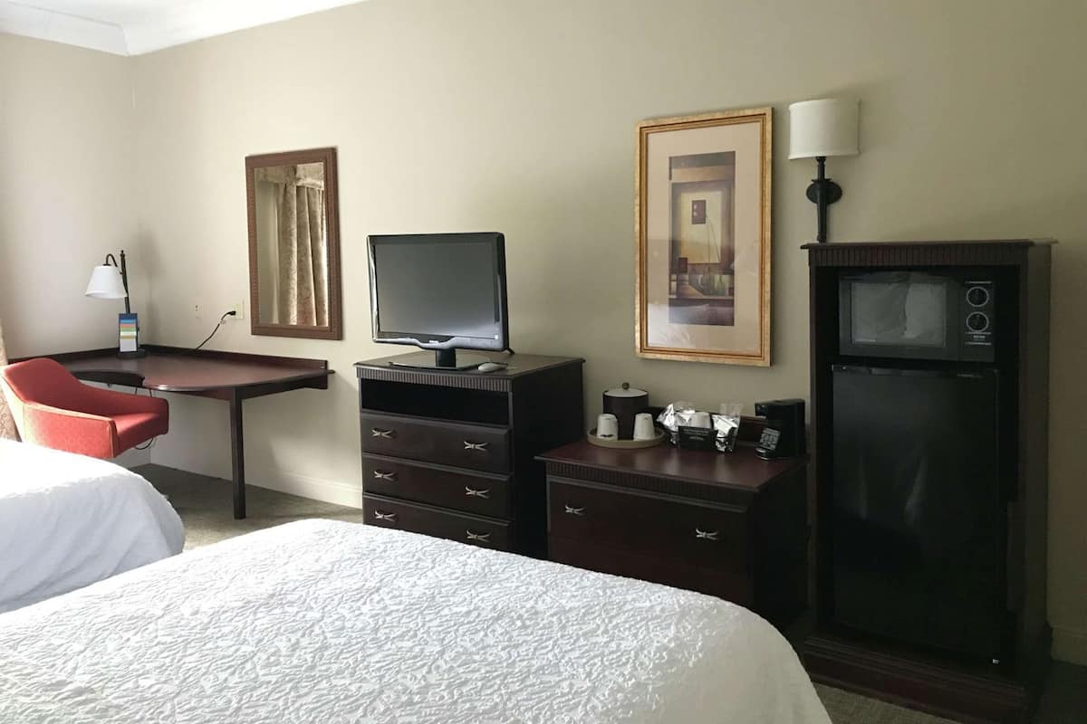 A hotel room with a bed, a desk with a computer, a chair, a dresser, a refrigerator, and two framed pictures on the wall.