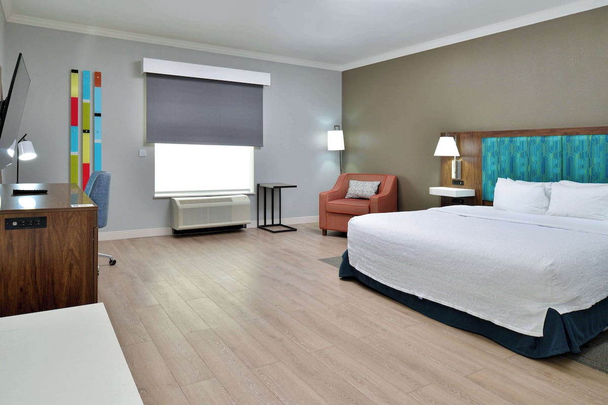 A hotel room with a bed, a desk, a chair, a lamp, a window with blinds, and a wall-mounted air conditioner.