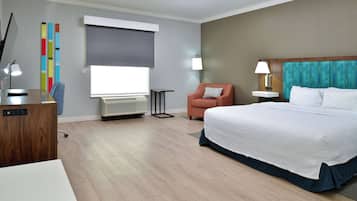 Ein Hotelzimmer mit einem Bett, einem Schreibtisch, einem Stuhl, einer Lampe, einem Fenster mit Jalousien und einer Wandklimaanlage.
