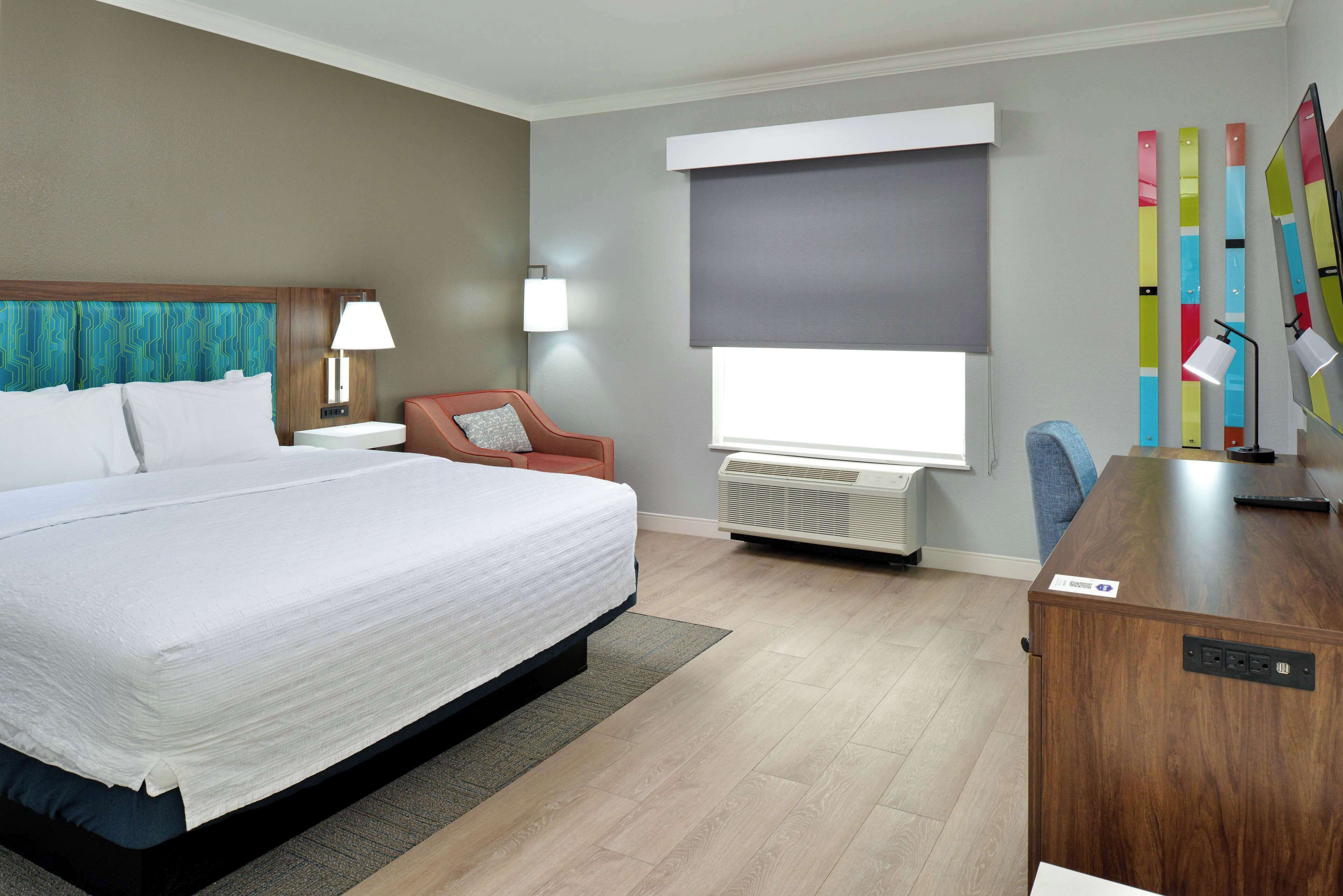 Habitación de hotel con una cama grande, un escritorio, una silla, una lámpara, un calentador y una ventana con persianas.
