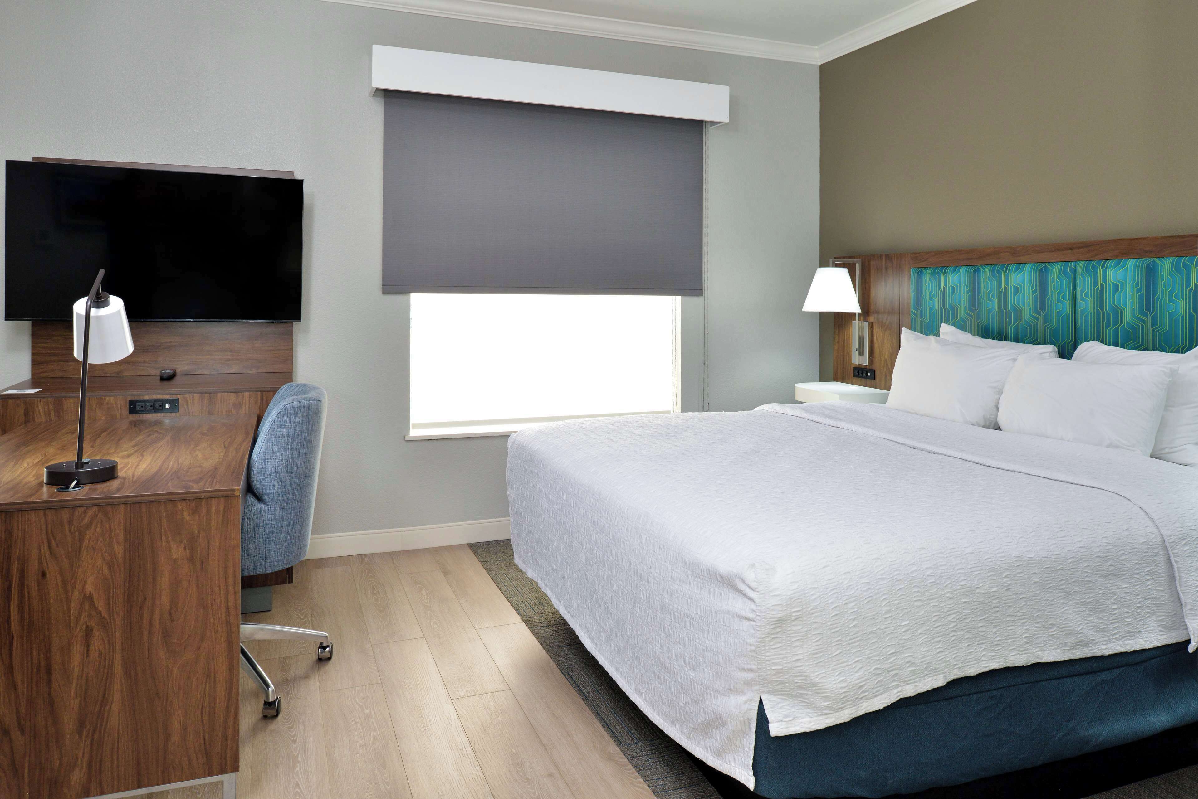 A hotel room with a bed, a desk with a TV, a chair, a window with a blind, and a nightstand with a lamp.