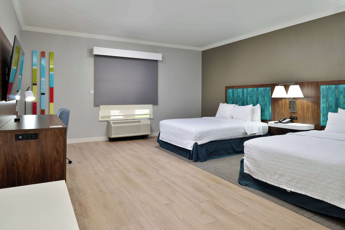 A hotel room with two beds, a desk, a chair, a desk lamp, a telephone, and a wall-mounted air conditioner.