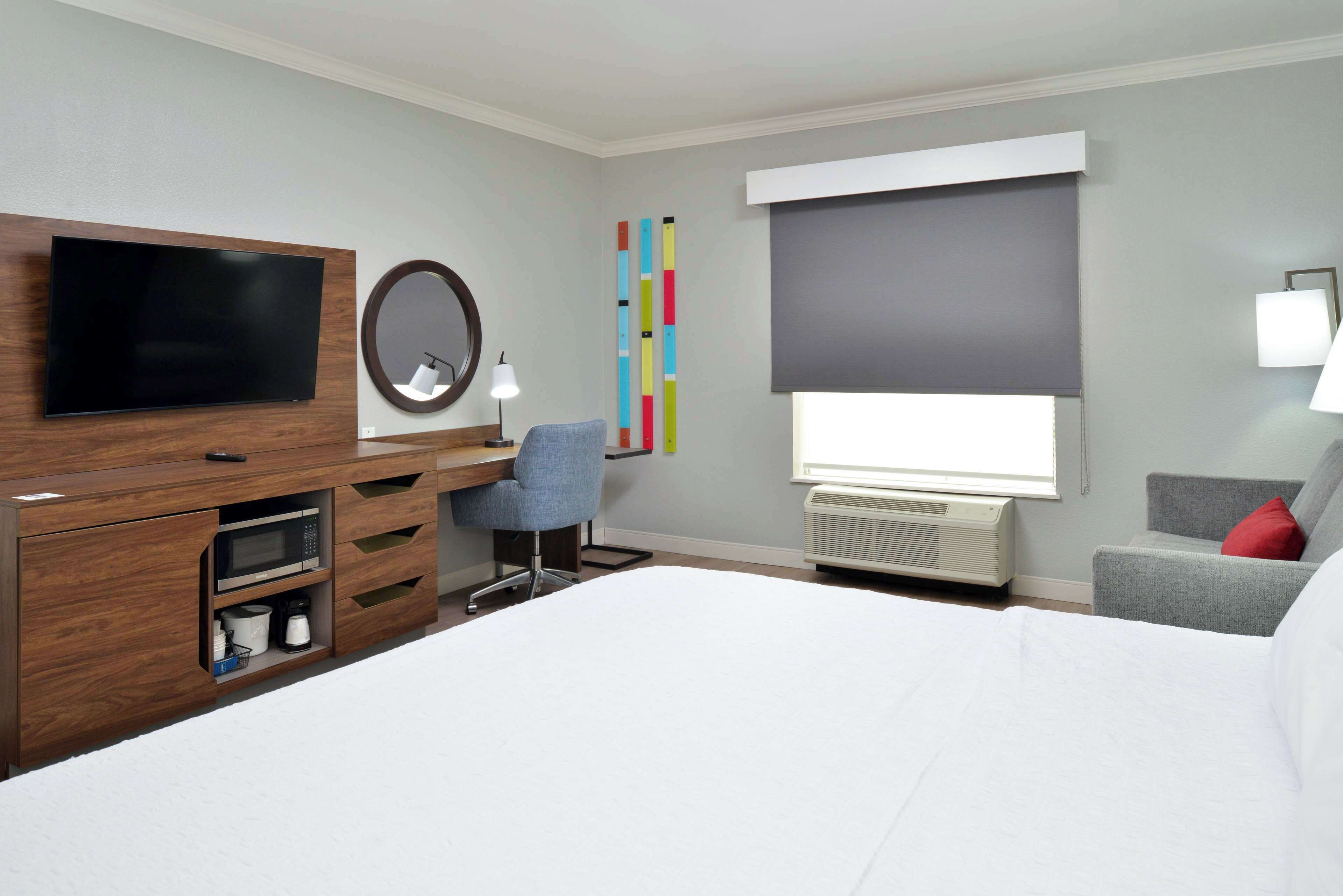 A hotel room with a bed, a desk with a chair, a flat-screen TV, a mirror, a window with blinds, and a wall-mounted air conditioner.