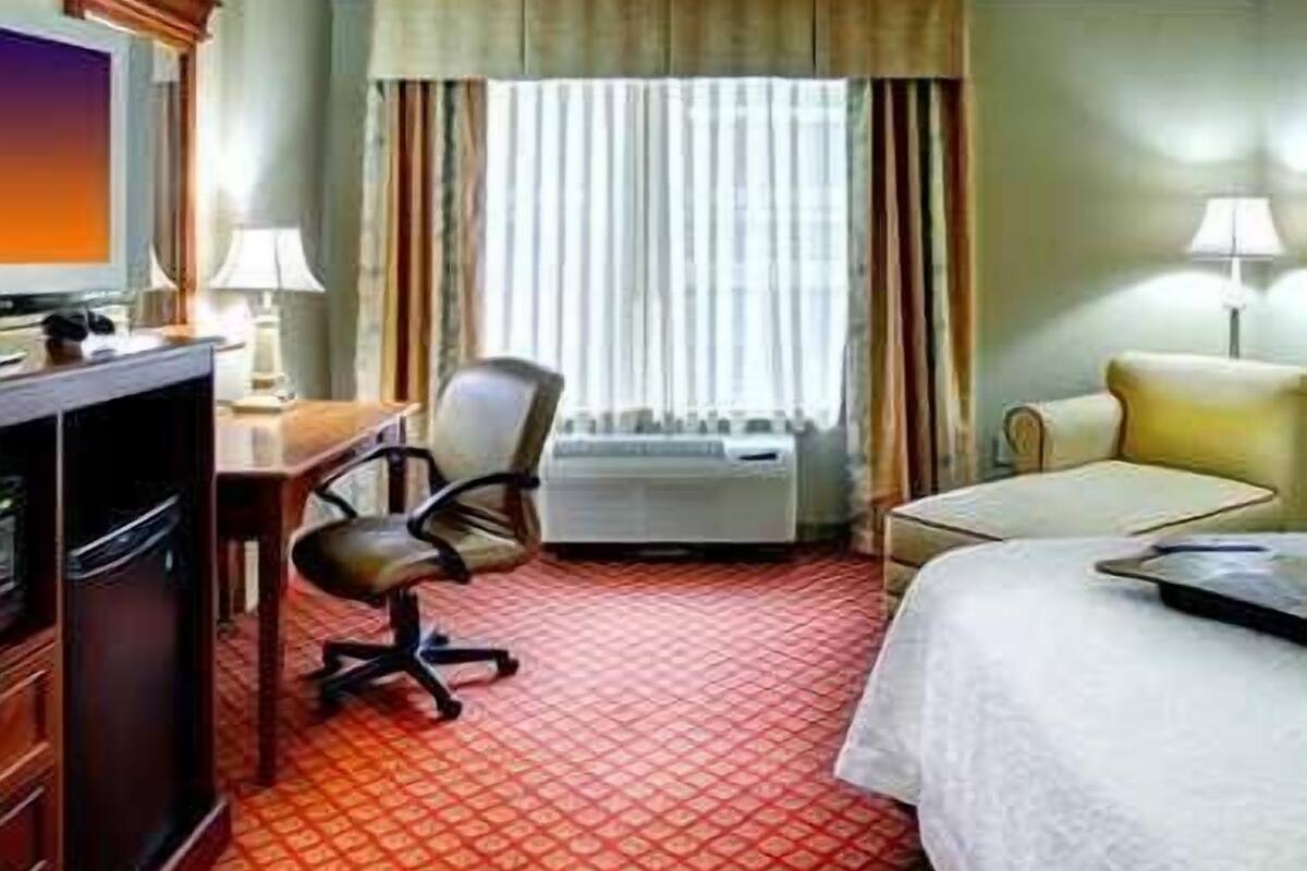 A hotel room with a bed, a desk with a chair, a television, and a window with curtains.