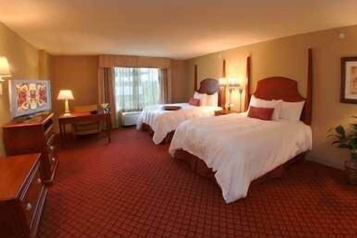 A hotel room with two beds, a desk, a chair, a lamp, and a window.