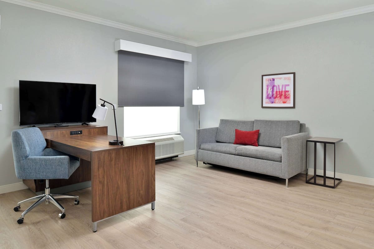 A modern living room with a wooden desk, a grey sofa, a flat-screen TV, and a framed artwork on the wall.