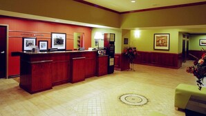 A hotel reception area with a wooden reception desk, framed pictures on the wall, a flower arrangement, and a green upholstered bench. - Hampton Inn & Suites Greenville (Greenville)