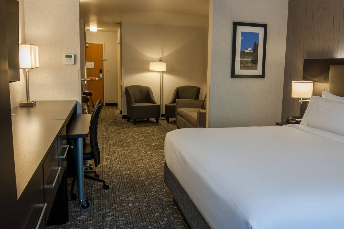 A hotel room with a large bed, a desk with a chair, two armchairs, a nightstand with a lamp, and a framed picture on the wall.
