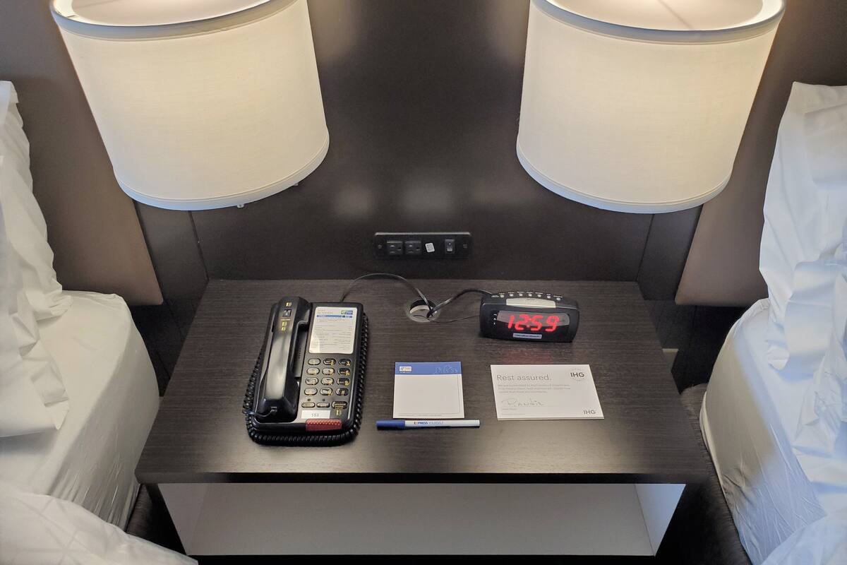 A hotel room with two beds, a bedside table with a phone, a digital clock, a pen, and a card.