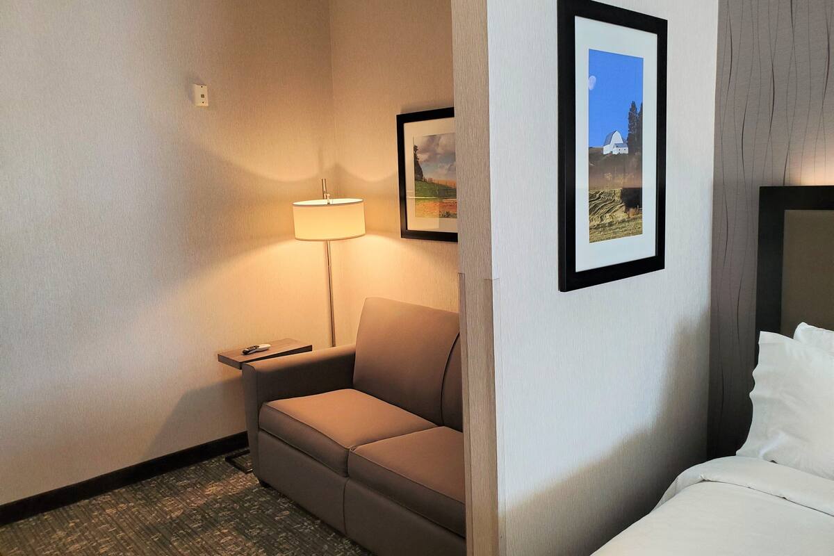 A hotel room with a bed, a sofa, a lamp, and two framed pictures on the wall.