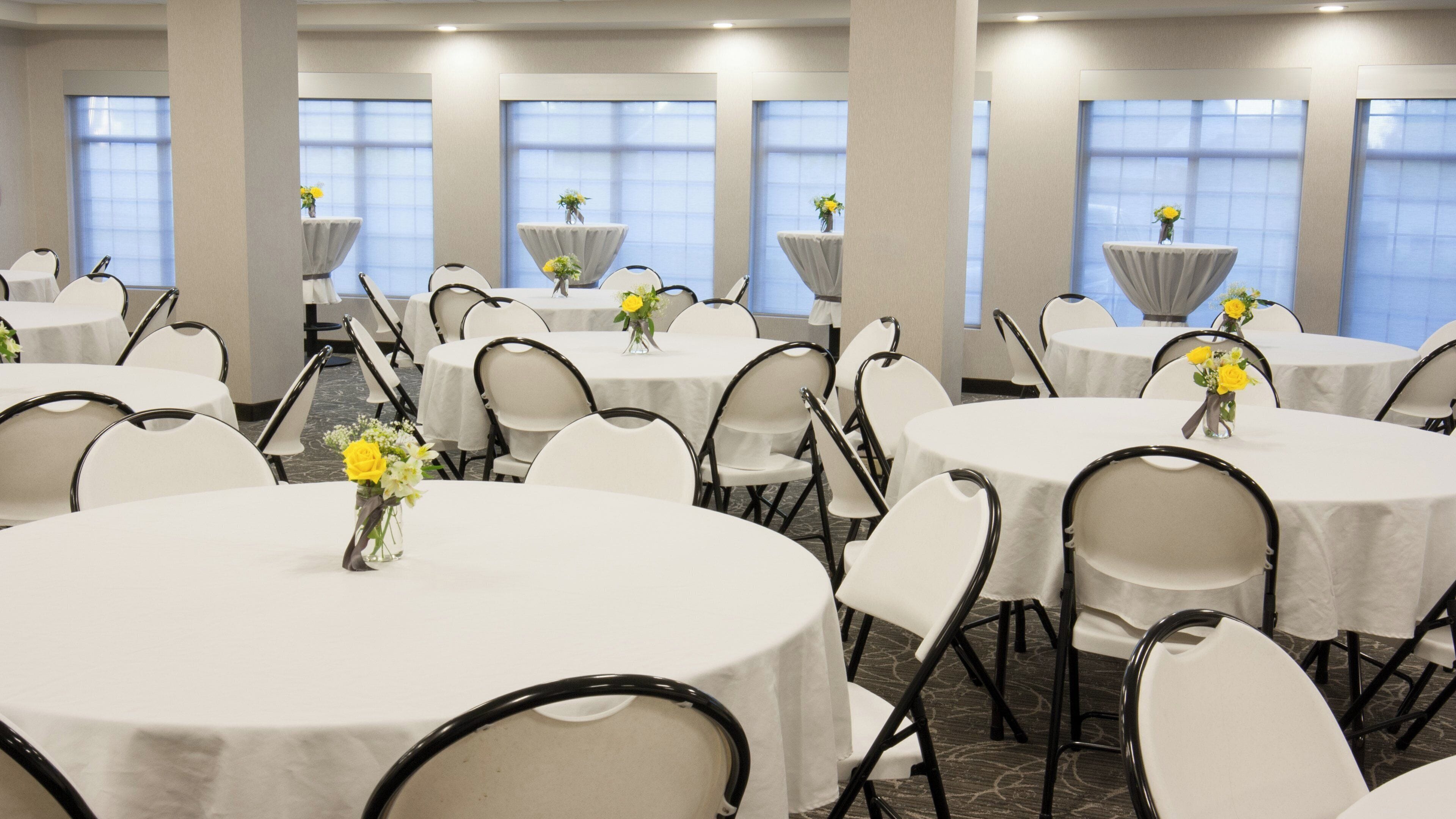 A hotel conference room with round tables, white chairs, and floral centerpieces.