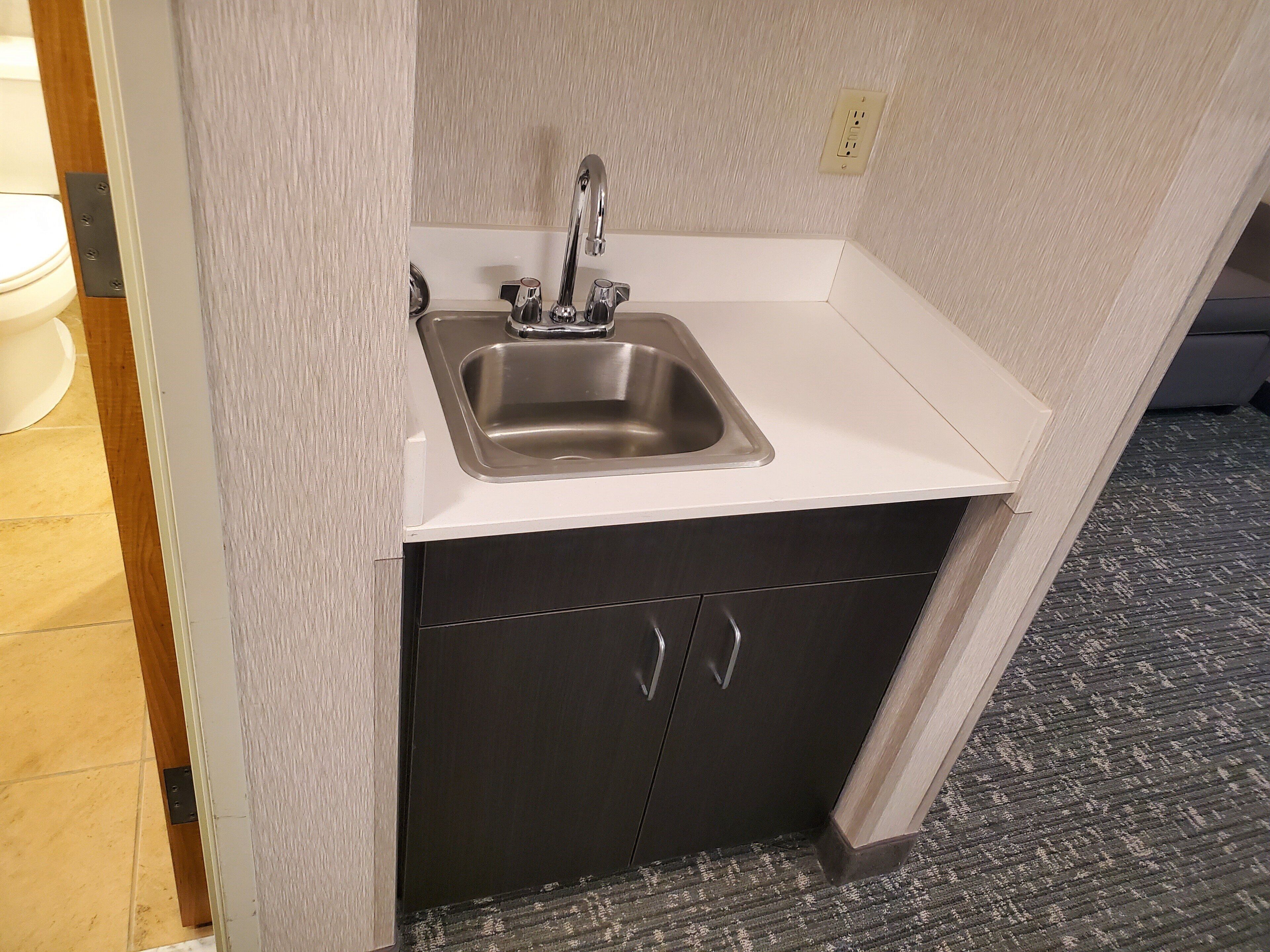 A small kitchenette with a stainless steel sink, a dark cabinet, and a white countertop.