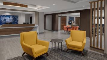 A modern hotel lobby with two yellow armchairs, a reception desk, and a dining area in the background.