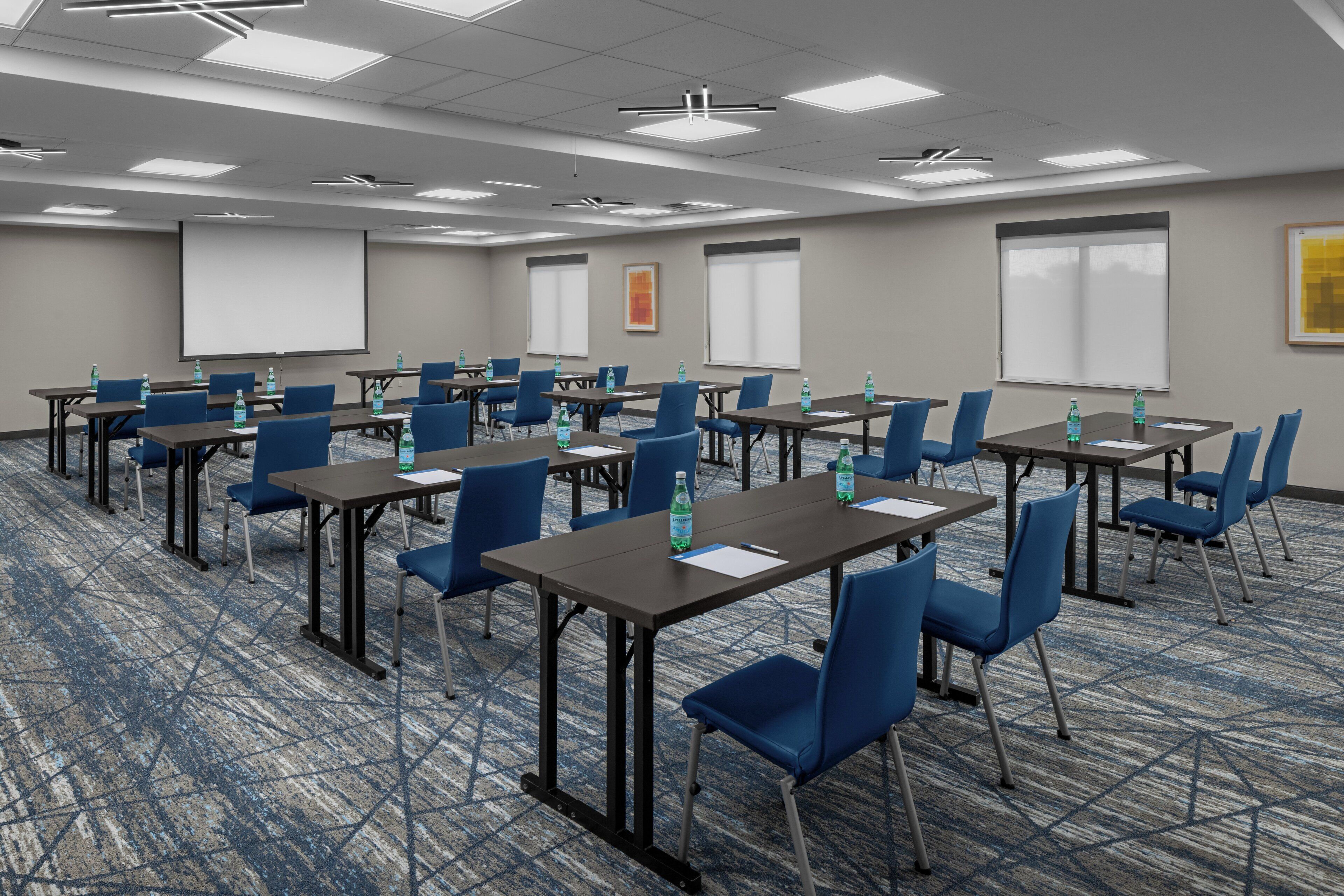 Sala de conferências com várias mesas e cadeiras, equipada com garrafas de água e ecrãs de apresentação.
