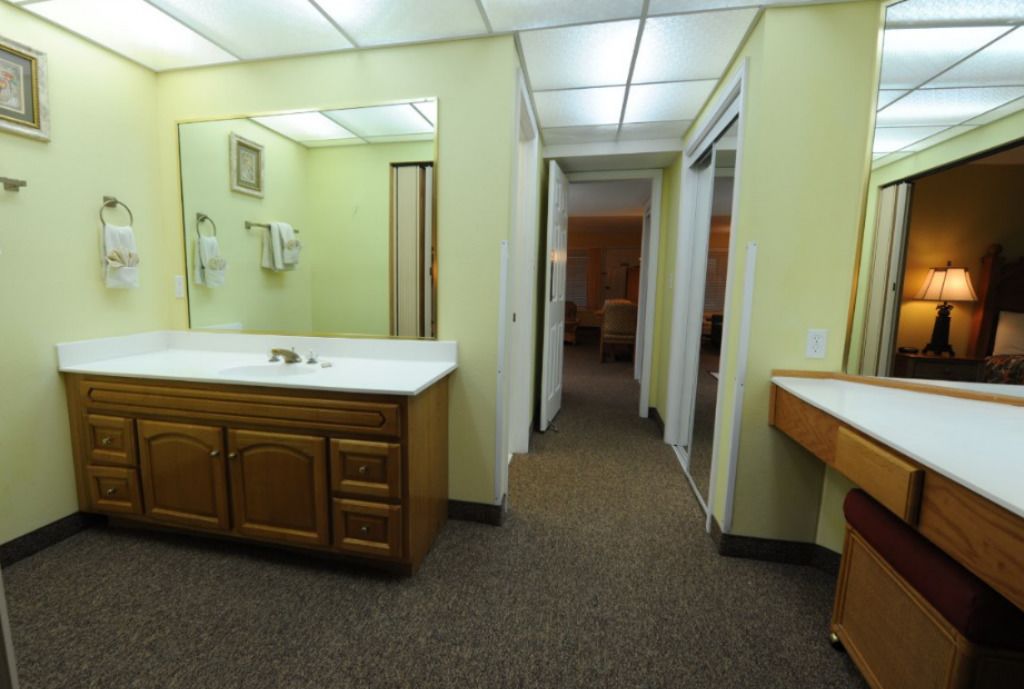 A hotel bathroom with a large vanity, a mirror, and a sink. There is a hallway leading to another room.