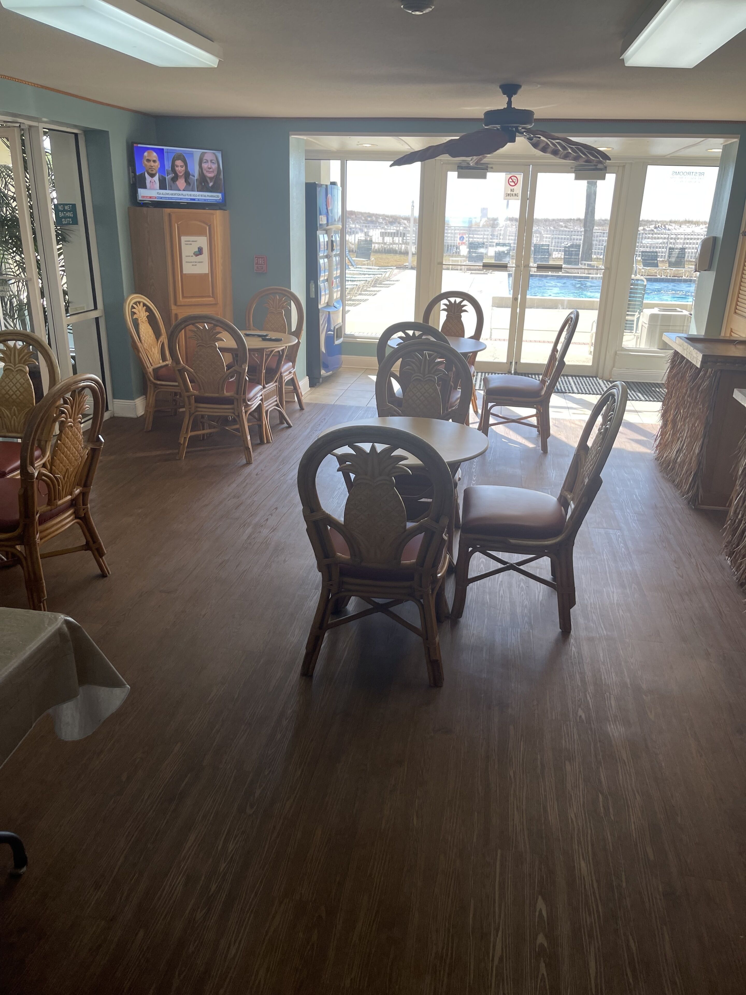 A dining area with wooden chairs and tables, a television mounted on the wall, and large windows offering a view of the city.