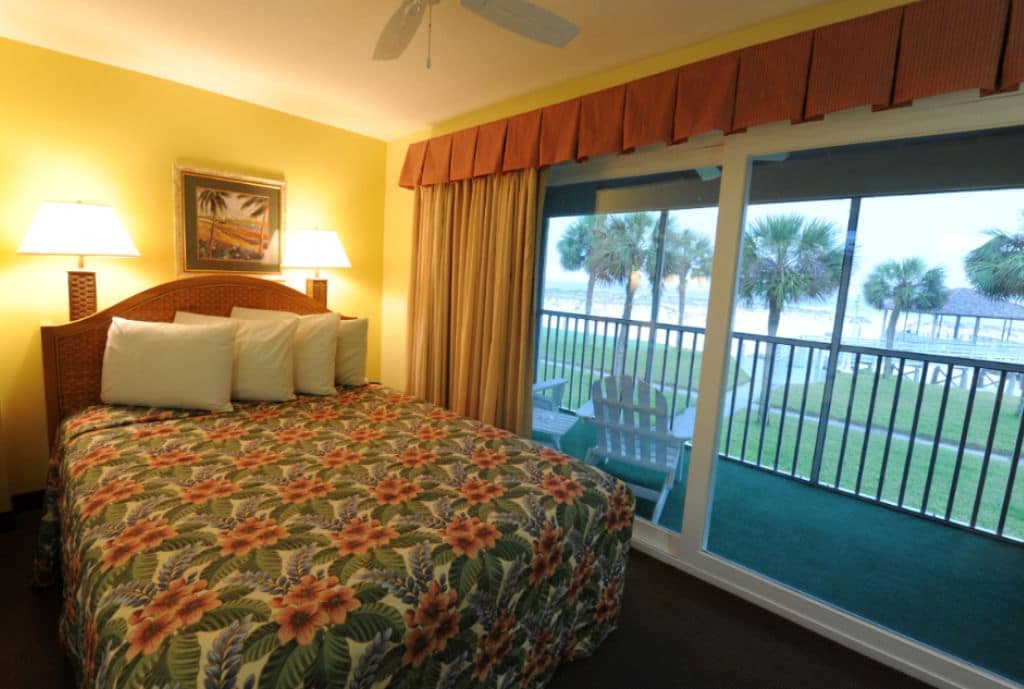 A hotel room with a bed, two lamps, a painting, and a balcony with a view of palm trees and a beach.