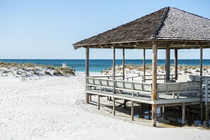 A beach gazebo with a wooden deck and a sloped roof, situated on a sandy beach with the ocean in the background.